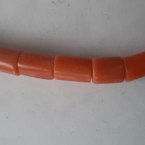 May include: Close-up of a necklace featuring coral-colored, cylindrical beads. The beads are evenly sized and strung together, creating a curved line against a white background. The necklace appears to be made of a smooth, polished material.