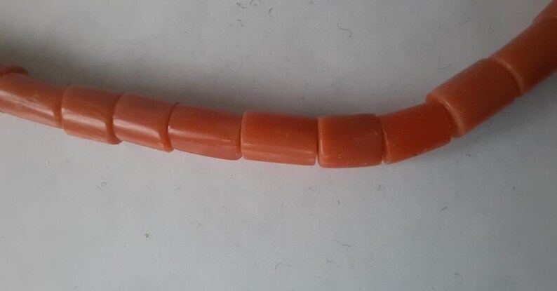 May include: Close-up of a necklace featuring coral-colored, cylindrical beads. The beads are evenly sized and strung together, creating a curved line against a white background. The necklace appears to be made of a smooth, polished material.