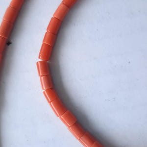 May include: Close-up of a necklace made of bright orange, rectangular beads. The beads are strung together, forming a curved shape against a white background. The necklace appears to be a jewelry item.