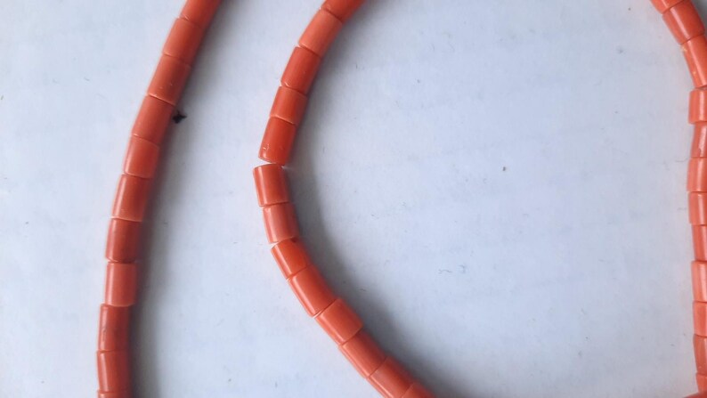 May include: Close-up of a necklace made of bright orange, rectangular beads. The beads are strung together, forming a curved shape against a white background. The necklace appears to be a jewelry item.