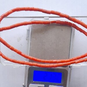 May include: A necklace made of small, rectangular, orange beads. The necklace is draped over a clear plastic container, which is on a digital scale. The scale's display shows a blue light.