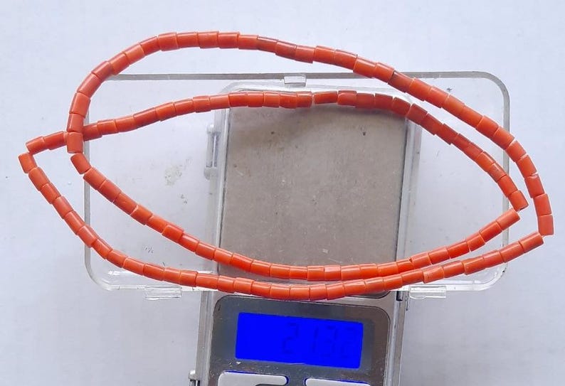 May include: A necklace made of small, rectangular, orange beads. The necklace is draped over a clear plastic container, which is on a digital scale. The scale's display shows a blue light.