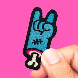 Op de afbeelding: Geborduurde patch met een blauwe hand die het rock-and-roll-teken maakt, met een zwarte omtrek. De patch heeft een roze mond en een botachtig detail. De achtergrond is effen roze.