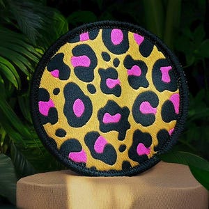 Op de afbeelding: Een ronde, zwart omrande patch met een luipaardprint. De print heeft zwarte vlekken op een gele achtergrond met roze accenten.