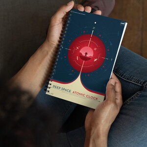 May include: A blue and red spiral bound notebook with the text "Deep Space Atomic Clock" and a graphic of a clock face with a red circle in the center. The clock face has numbers 1 through 12 and a small figure of an astronaut standing on a red planet.
