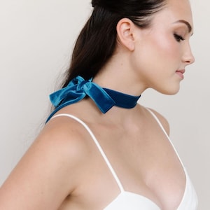 May include: A woman wearing a white spaghetti strap top with a teal blue velvet choker tied in a bow at the back of her neck.