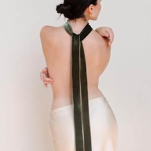 May include: A dark green velvet scarf is draped over the shoulders and hangs down the back. The scarf is a deep, rich green color. The model is wearing a cream-colored skirt. The background is a plain, light beige color.
