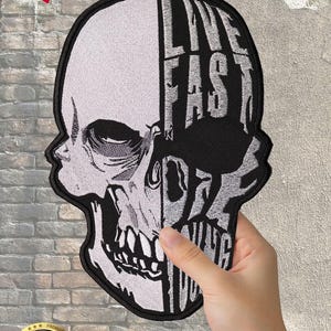 May include: A black and white skull patch with the words "LIVE FAST DIE YOUNG" in a bold, stylized font. The patch is held by a hand, and the design is split vertically. A "NEW" banner and a "PREMIUM QUALITY" sticker are in the image.