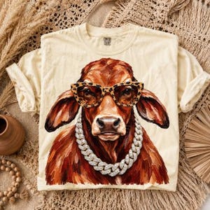 May include: A cream-colored short-sleeve t-shirt featuring a graphic of a brown cow wearing leopard-print glasses and a thick silver chain necklace. The shirt has a crew neckline and a Comfort Colors brand label printed on the inside neck area.