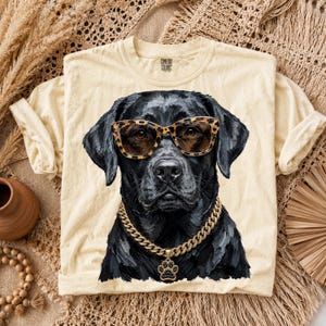 May include: A cream-colored t-shirt featuring a graphic of a black Labrador dog wearing leopard print glasses and a gold chain necklace with a paw print pendant. The shirt has a crew neckline and a Comfort Colors brand label on the inner collar.