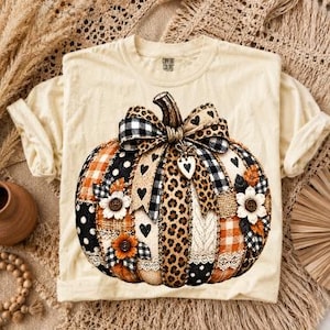 Puede incluir: Una camiseta de color crema con un gran gráfico de una calabaza decorada con varios patrones, incluyendo estampado de leopardo, cuadros vichy en blanco y negro, cuadros naranjas y lunares, acentuada con flores de tela y un dije de corazón.