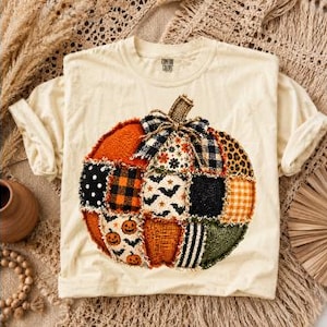 May include: A cream-coloured t-shirt featuring a large, central graphic of a pumpkin constructed from various fabric patches. The pumpkin design includes patterns like black and white bats, orange plaid, leopard print, and polka dots, topped with a plaid bow.