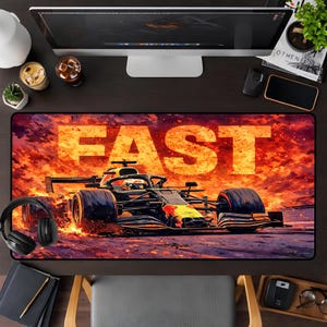 May include: A rectangular desk mat featuring a Formula 1 race car design with the word "FAST" in large, bold letters. The background is a fiery orange and red, with the car in shades of black, yellow, and red.