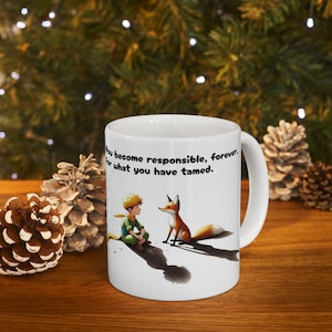 May include: White ceramic mug featuring an illustration of the Little Prince and a fox, with the quote "You become responsible, forever, for what you have tamed." The mug has a white handle and is set against a blurred background.