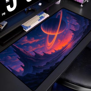 Può includere: Tappetino per scrivania rettangolare con un design a tema spaziale. L'opera d'arte raffigura un pianeta con anelli sullo sfondo di un tramonto infuocato e un terreno montuoso. Sono visibili una tastiera e un monitor.
