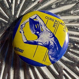 May include: A round button with a blue border and a yellow center. The center features a blue illustration of a person with the words "ORG ASM ADDICT" and "ANZCOCKS".