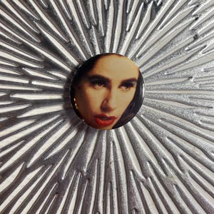 May include: A round button pin featuring a portrait of a person with dark hair, dark eyes, and red lipstick. The pin is set against a silver, textured, radial background.