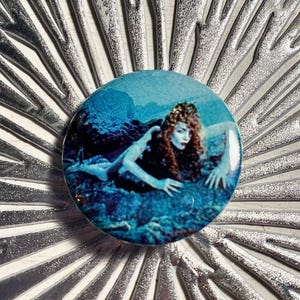 May include: A round button with a vintage image of a woman in a blue underwater scene. The button is set against a silver, starburst-like background. The woman has red hair and is reaching out.