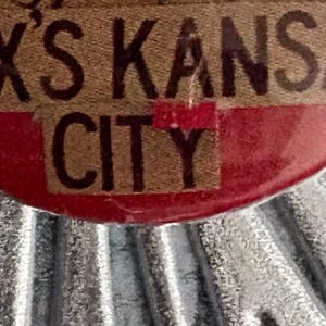 May include: Close-up of a brown and red label with the text "AX'S KANSAS CITY" in a bold, vintage font. The label has a textured appearance and is set against a corrugated metal background.