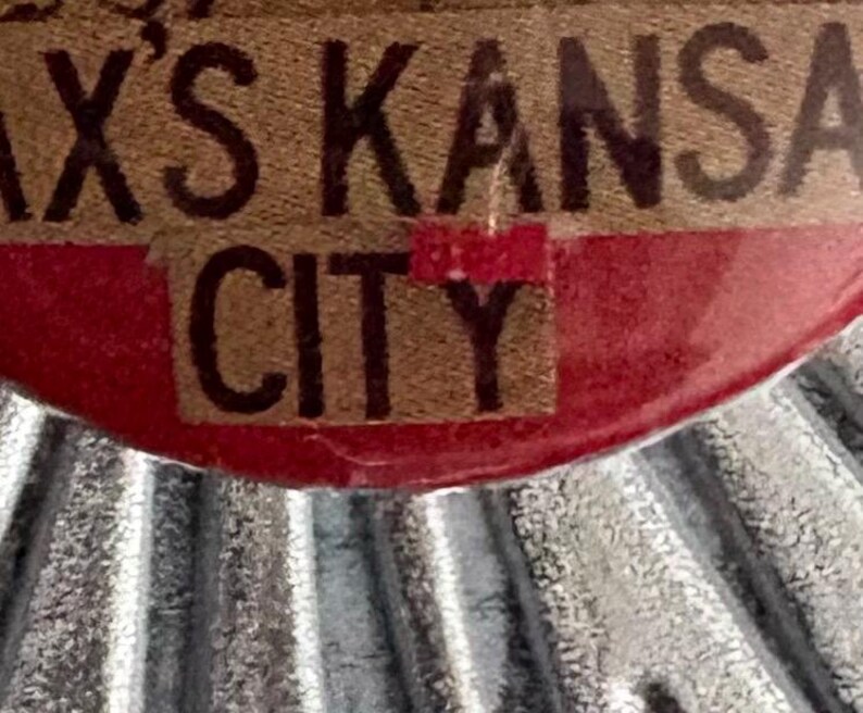 May include: Close-up of a brown and red label with the text "AX'S KANSAS CITY" in a bold, vintage font. The label has a textured appearance and is set against a corrugated metal background.