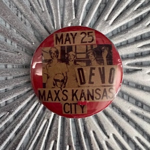 May include: A red button with the text "MAY 25" at the top, and "DEVO" and "MAX'S KANSAS CITY" in the centre. The button features a sepia-toned image of a band. The button is round.