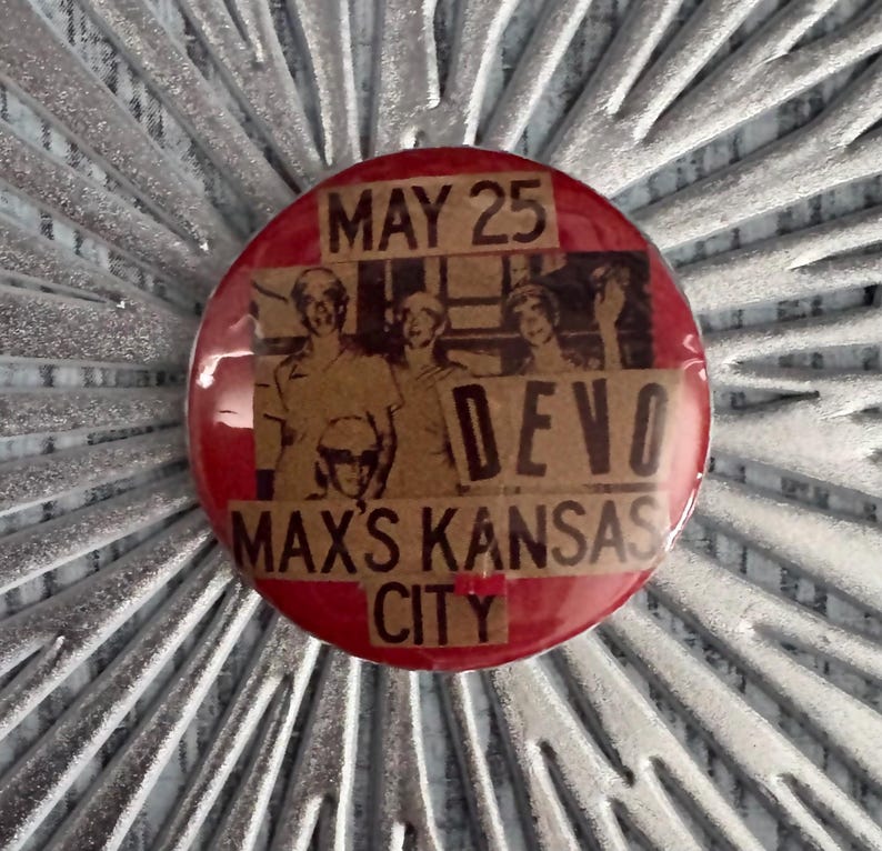 May include: A red button with the text "MAY 25" at the top, and "DEVO" and "MAX'S KANSAS CITY" in the centre. The button features a sepia-toned image of a band. The button is round.