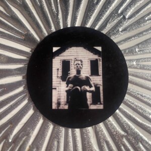 May include: A black, circular button featuring a black and white photograph of a person standing in front of a house. The button is set against a silver, textured background with a radial pattern.