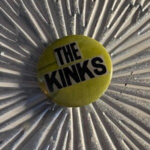 May include: A round, yellow button with the text "THE KINKS" in black block letters. The button is set against a silver, starburst-patterned background. A music-themed collectible.