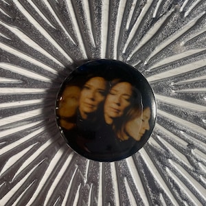 May include: A round, black button featuring a sepia-toned image of a person's face in multiple exposures. The button is set against a textured, silver-colored background with radial lines.