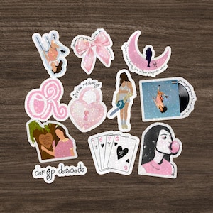 May include: A collection of pink and white stickers with various designs, including a bow, a crescent moon, playing cards, and a girl blowing a bubble. Some stickers have text, such as "Olivia Rodrigo" and "drop dead".