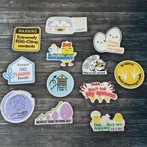 May include: A selection of humorous stickers with egg and chick illustrations. The stickers display phrases such as "Egg Math is Different" and "Breakfast Bonds Maturing Daily." The stickers are arranged on a dark wooden background.