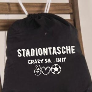 May include: Black drawstring bag with white text reading "STADIONTASCHE" above "CRAZY SH... IN IT" and a peace sign, heart, and football graphic. The bag has white drawstrings and is on a wooden chair.