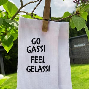 Könnte beinhalten: Weiße Socken mit dem Text "GO GASSI FEEL GELASSI" in Schwarz, hängen an einer Holzklammer an einem Ast. Der Hintergrund zeigt grüne Blätter und eine verschwommene Außenansicht.