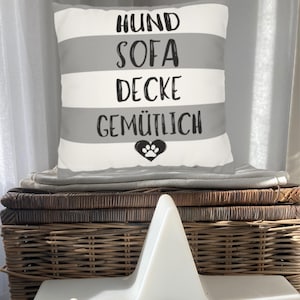 Könnte beinhalten: Ein dekoratives Kissen mit grauen und weißen Querstreifen. Es trägt die schwarzen Worte "HUND SOFA DECKE GEMÜTLICH" sowie ein Pfotenabdruck-Herz-Design. Das Kissen liegt auf einem Stapel Decken.