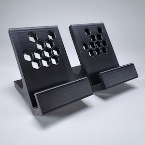 May include: Two black 3D printed phone stands with a honeycomb design. The stands are angled to hold a phone upright.