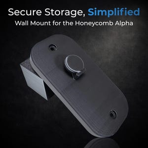 May include: A black wall mount for the Honeycomb Alpha, with the text "Secure Storage, Simplified". The mount has a rounded rectangular shape with a circular knob and two holes. It is resting on a gray, angled support.