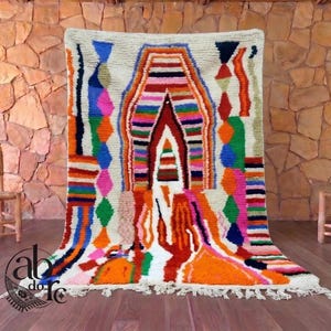 May include: A rectangular area rug with a cream-coloured base featuring a central arch motif and vertical geometric patterns in vibrant shades of orange, pink, blue, green, and black. The rug has a thick pile texture and white fringe at the bottom.