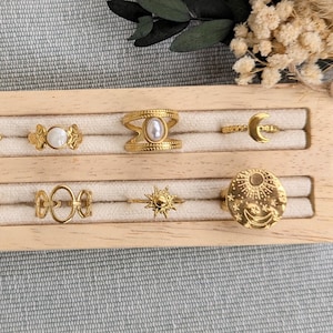Peut inclure: Une collection de bagues dorées exposées sur un présentoir en bois. Les bagues présentent divers motifs, notamment des formes ouvertes, des détails floraux, des perles et des motifs célestes comme un soleil et une lune. Les bagues sont présentées sur un tissu clair.