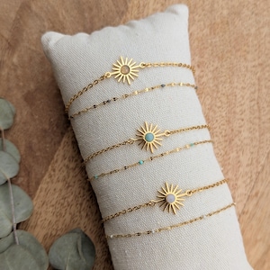 Op de afbeelding: Gouden kettingarmbanden met zonneparels, elk met een andere edelsteen. De armbanden worden gepresenteerd op een wit kussen tegen een houten achtergrond. De edelstenen zijn lichtbruin, turkoois en wit.