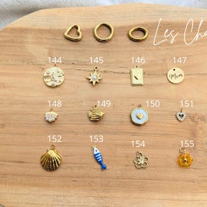 May include: A collection of gold-toned metal jewelry charms labeled 144 through 155, including shapes like a moon, star, heart, fish, shell, and flower. The text Les Charms is written in the upper right corner above the numbered metal charms.