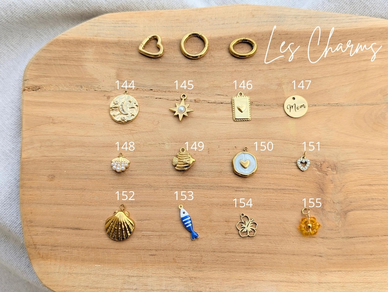 May include: A collection of gold-toned metal jewelry charms labeled 144 through 155, including shapes like a moon, star, heart, fish, shell, and flower. The text Les Charms is written in the upper right corner above the numbered metal charms.