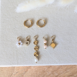 Puede incluir: Una colección de pendientes dorados, que incluye aros pequeños y pendientes con colgantes. Los colgantes presentan una flor con cuentas blancas, un corazón con piedras transparentes, una cadena vertical de discos dorados, perlas rosas y un diseño de estrella. Se muestran sobre una superficie gris claro.