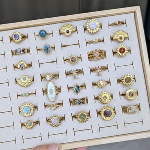 Pode incluir: Uma coleção de anéis dourados exibidos numa bandeja de veludo claro. Os anéis apresentam vários designs, incluindo motivos de raios de sol, formas geométricas e pedras incrustadas em cores como azul, vermelho, verde, branco e castanho.