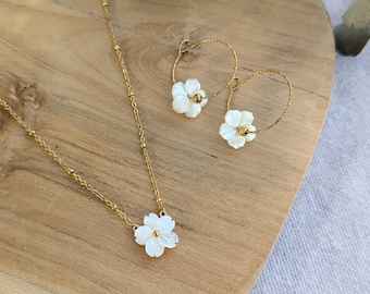 May include: A gold chain necklace featuring a small white flower pendant with a central gold bead, accompanied by a pair of gold hoop earrings, each adorned with a matching white flower charm. The jewelry set displays a delicate, polished finish.