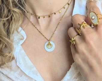 May include: A gold-toned layered necklace set featuring a delicate chain with small teardrop charms and a second chain with a circular white shell pendant displaying a gold seashell cutout. Three gold rings are worn, including two starfish designs and one oval ring with a white center stone.