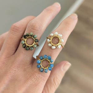 Peut inclure: Trois bagues dorées, chacune avec un motif floral. Une bague a des perles vertes et noires, une autre des perles blanches et la troisième des perles bleues. Les bagues sont présentées sur des doigts sur un fond neutre.