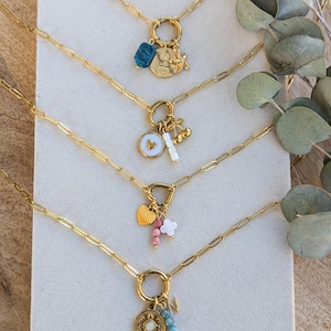 May include: Four gold-toned chain necklaces featuring paperclip links and circular clasps. Each necklace displays a unique arrangement of charms, including a blue stone, a gold disc with heart engravings, a cherry charm, a white heart, and a letter N.