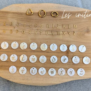 May include: A collection of jewelry components including gold-toned metal alphabet charms, three gold-toned metal rings in heart and circular shapes, and white circular mother-of-pearl charms featuring gold-toned script letters from A to Z.
