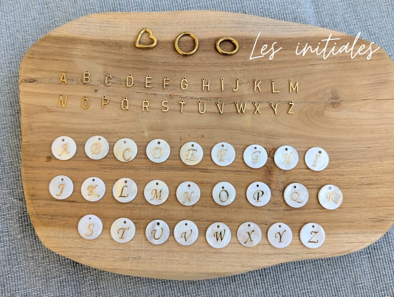 May include: A collection of jewelry components including gold-toned metal alphabet charms, three gold-toned metal rings in heart and circular shapes, and white circular mother-of-pearl charms featuring gold-toned script letters from A to Z.