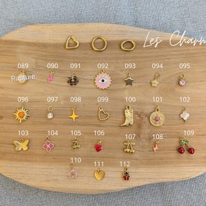 May include: A collection of gold-toned metal charms arranged on a wooden board, labeled with numbers 089 through 112. The charms include shapes like hearts, stars, flowers, a cowboy boot, an eye, cherries, and a lightning bolt, with some featuring enamel.